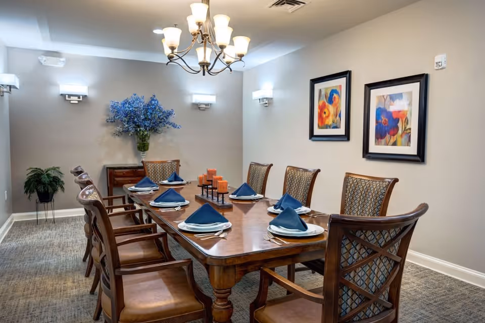 A formal dining room with a long wooden table set for eight people. Each place setting has a white plate, silverware, and a blue folded napkin. The room features a chandelier overhead, wall sconces for lighting, two framed floral paintings on the wall, a small side table with a large vase of blue flowers, and a potted plant in the corner.