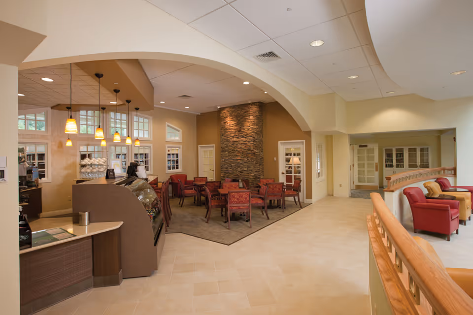 Interior view of a senior living facility lounge area with a small café counter on the left, pendant lights hanging from the ceiling, and several tables and chairs arranged on a carpeted section. The walls are painted in warm tones with a stone accent wall in the background. To the right, there are comfortable armchairs in red and tan colors near a wooden railing.