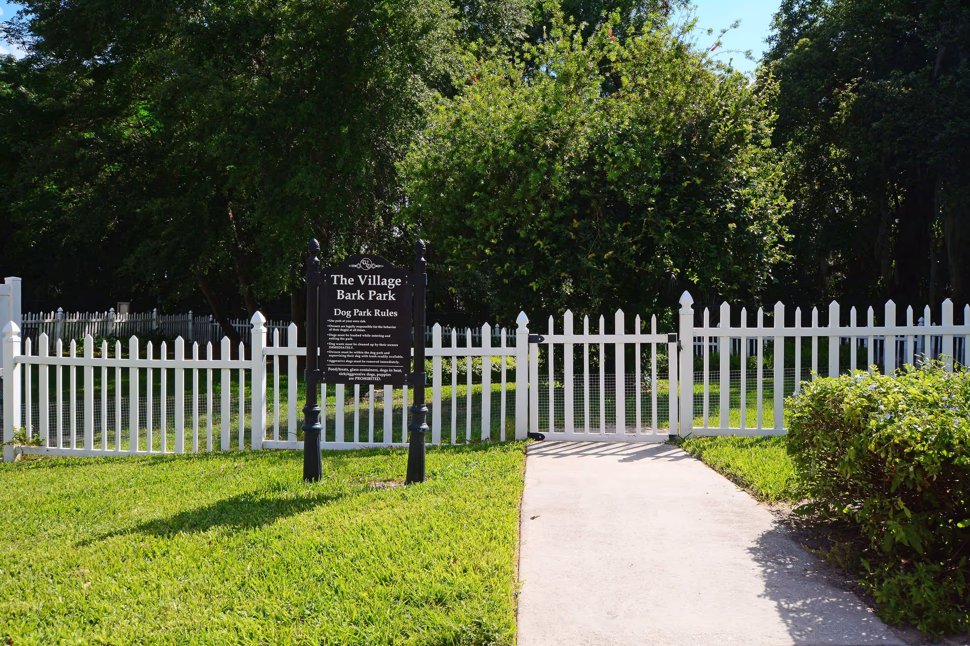 A white picket fence enclosing a grassy area with a black sign labeled 'The Village Bark Park Dog Park Rules' in front of the entrance gate. The area is surrounded by trees and bushes under a clear sky.