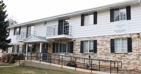 Exterior view of a two-story senior living facility building with white siding and brick facade, black shutters on windows, a small balcony, and a ramp leading to the entrance. A sign on the building reads 'Magnolia House by Adava Care'.