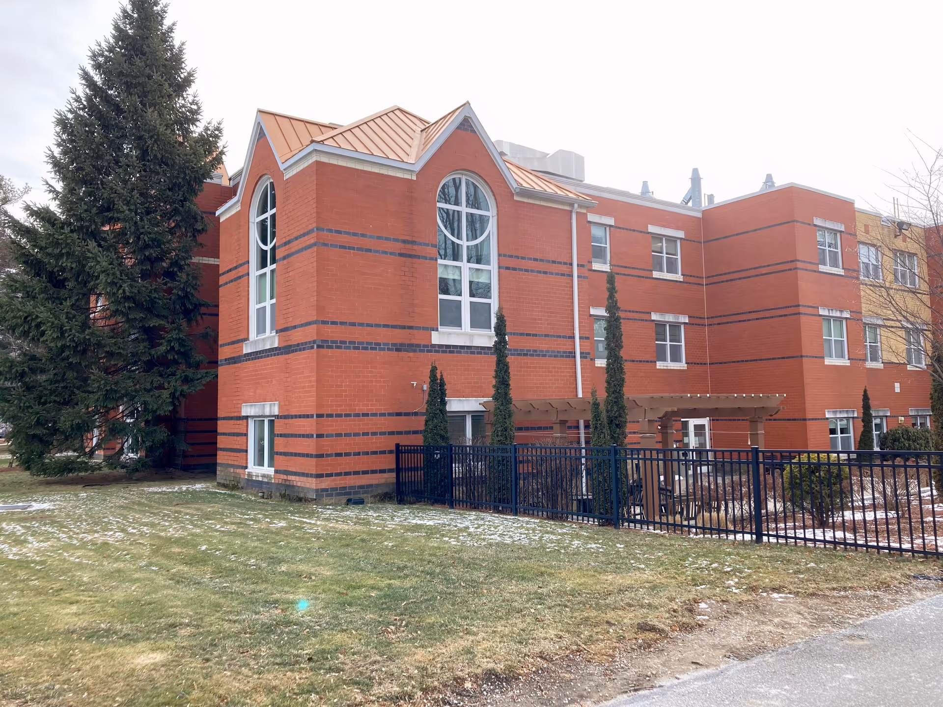 Exterior view of a red brick building with large arched windows and a copper-colored roof. There is a black metal fence enclosing a small garden area with a wooden pergola and some tall, narrow trees. A large evergreen tree is visible on the left side, and patches of snow are scattered on the grass and ground.