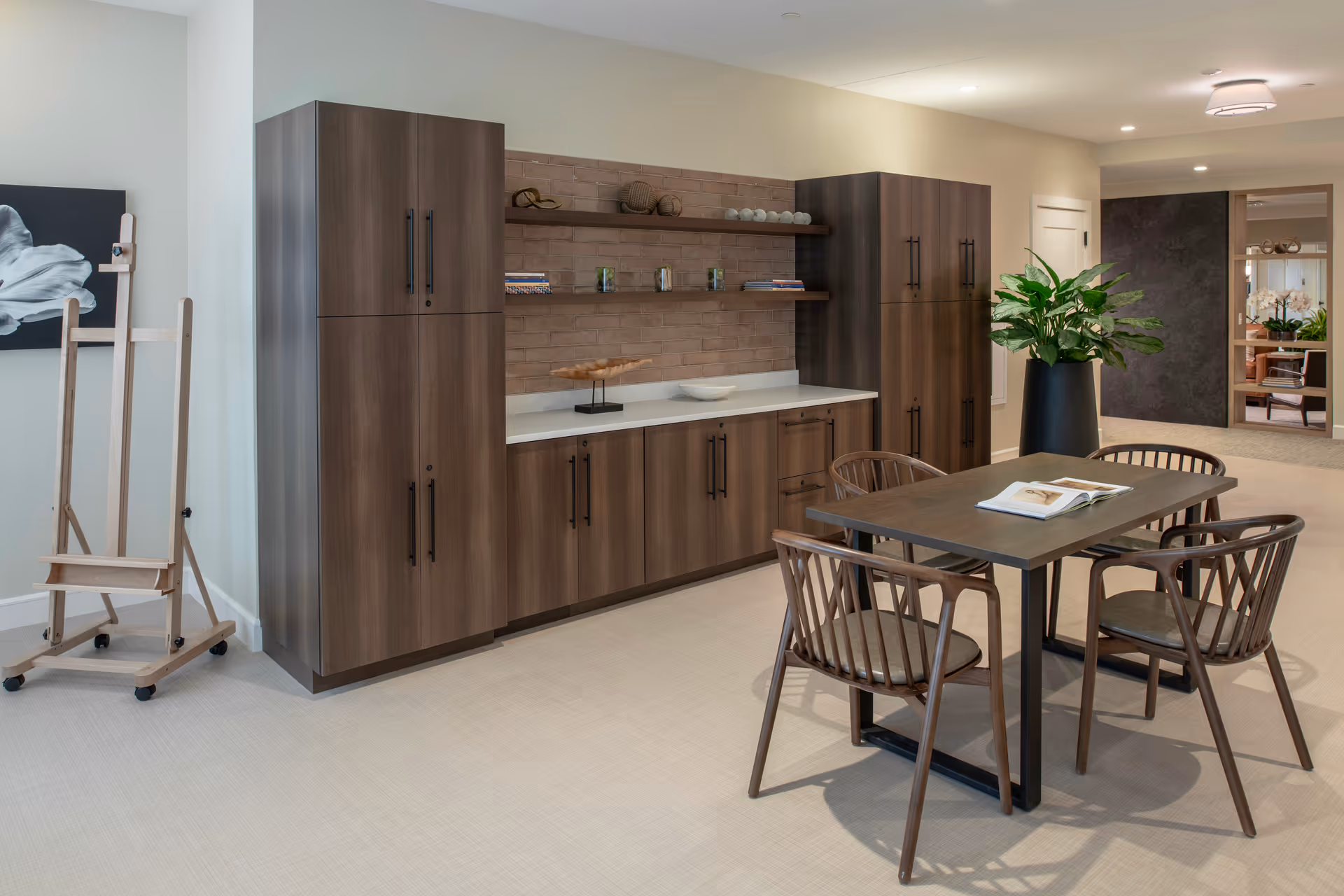 A modern interior space featuring a wooden table with four matching chairs, a large potted plant, and built-in wooden cabinets with shelves displaying decorative items. An easel with a black and white artwork is positioned on the left side, and a hallway with additional seating and plants is visible in the background.
