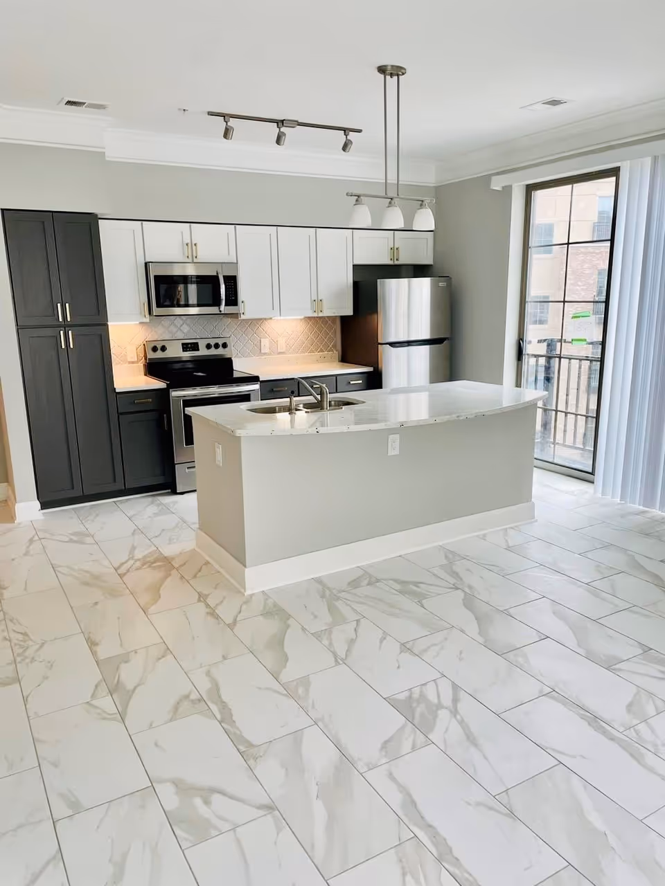 Modern kitchen with white and dark gray cabinets, stainless steel appliances including a refrigerator, stove, and microwave, a white marble countertop island with a sink, and large window with vertical blinds letting in natural light. The floor is covered with white tiles featuring gray marble patterns.