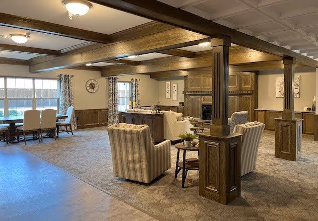 A spacious and well-lit living room area with wooden ceiling beams and columns. The room features comfortable seating including striped armchairs and a sofa arranged around a small table. Large windows with patterned curtains allow natural light to fill the space. The walls are decorated with framed artwork and a clock. The flooring is a combination of carpet and wood.