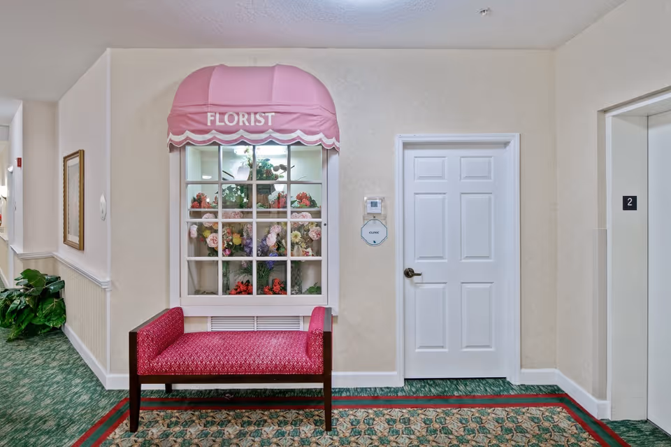 Indoor hallway featuring a pink 'FLORIST' window display of flowers, a red upholstered bench, a white door, and an elevator entrance.