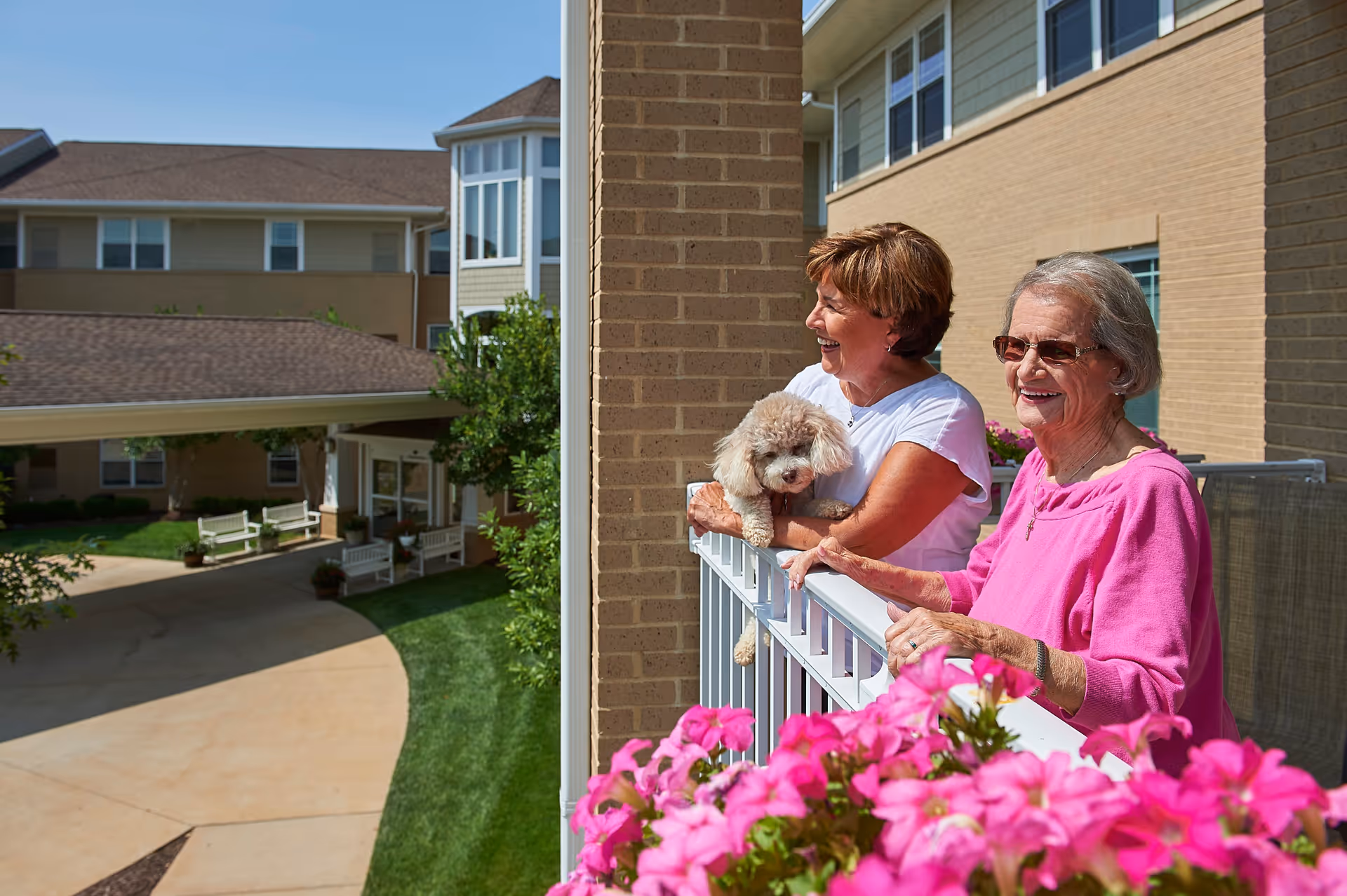 Two elderly women standing on a balcony of a senior living facility, smiling and enjoying the sunny day. One woman is holding a small fluffy dog. Pink flowers are in the foreground, and the building and courtyard are visible in the background.
