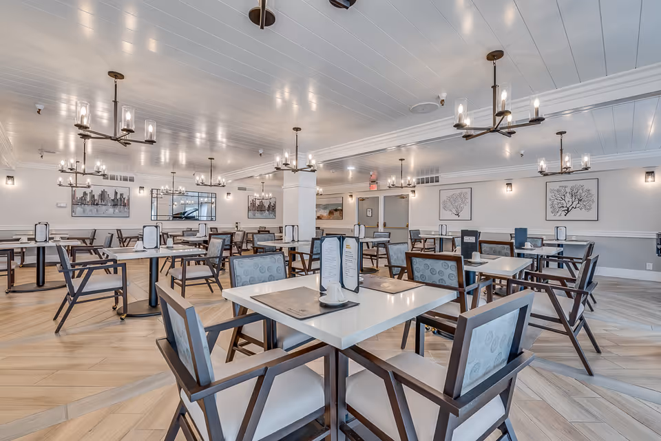 A spacious and well-lit dining room with multiple square tables and cushioned chairs arranged neatly. The room features modern chandeliers hanging from a white paneled ceiling, light wood flooring, and framed artwork on the walls. Menus and small condiment holders are placed on the tables.