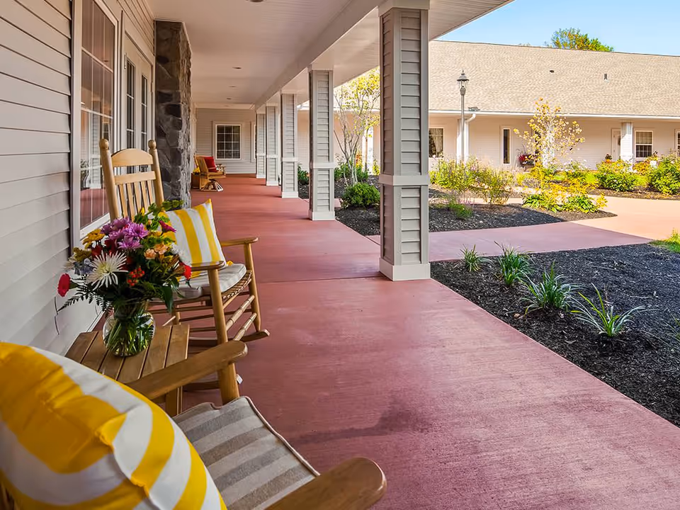 Covered outdoor walkway with wooden rocking chairs and small tables adorned with colorful cushions and a vase of flowers, leading to a building with beige siding and stone accents, surrounded by landscaped garden beds and a clear sky.