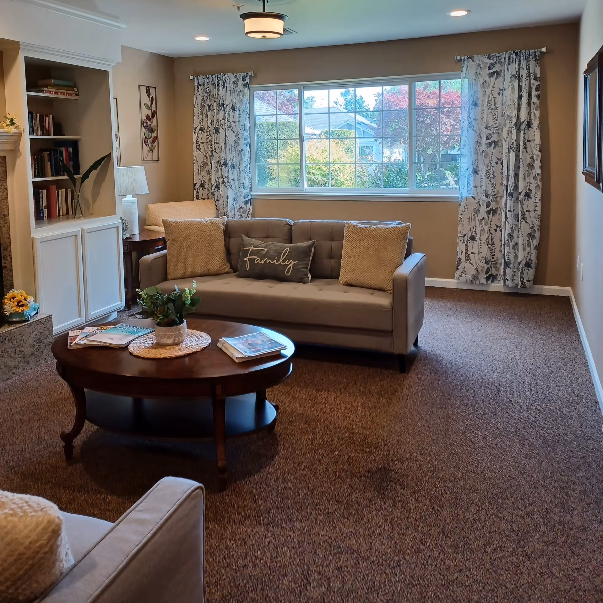 A cozy living room with a beige sofa adorned with three pillows, one of which has the word 'Family' embroidered on it. A wooden coffee table in front of the sofa holds a small potted plant and some magazines. To the left, there is a built-in bookshelf filled with books and a lamp on a side table next to an armchair. Large windows with patterned curtains allow natural light to brighten the room, and a carpeted floor covers the space.