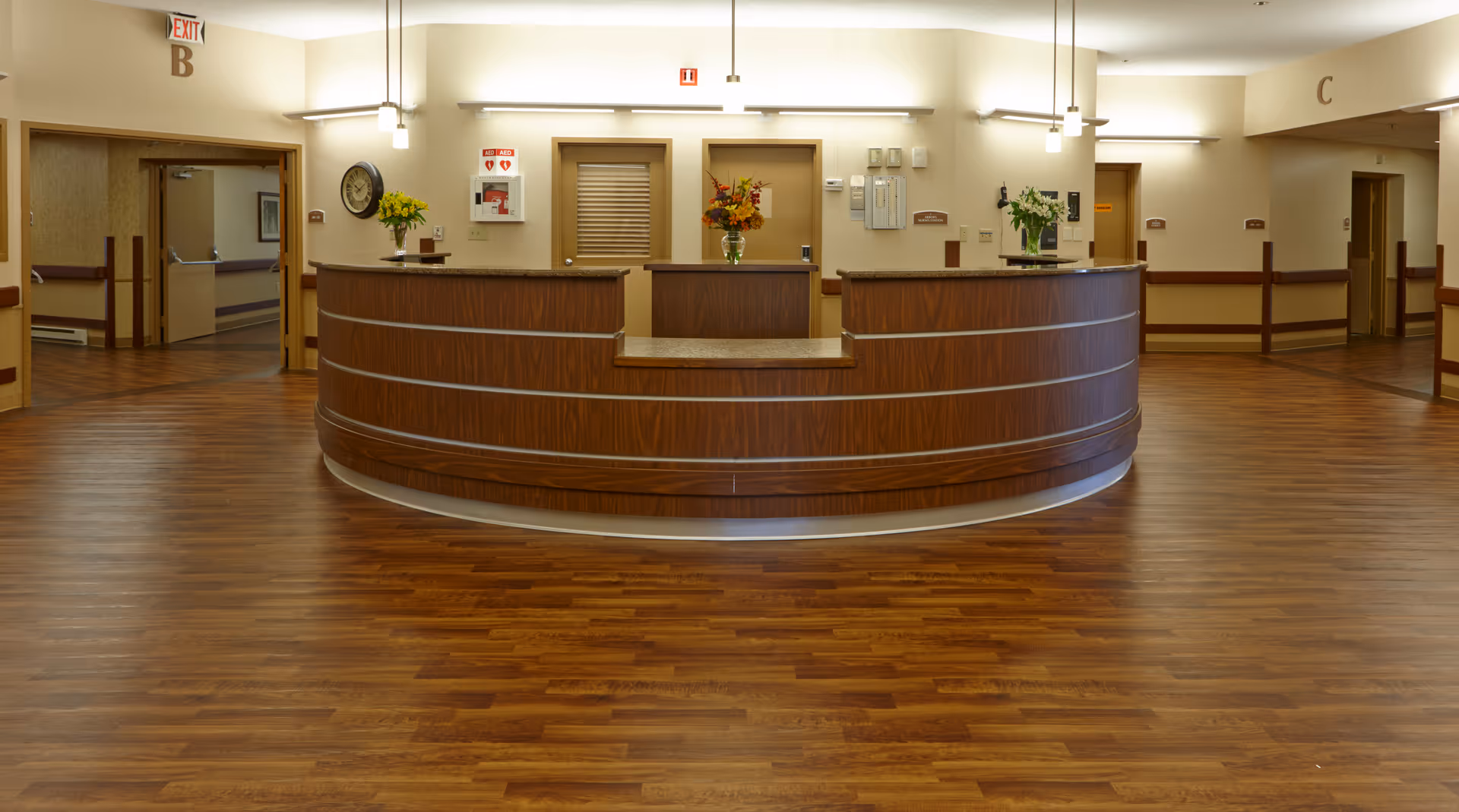A spacious reception area with a curved wooden front desk in the center. The floor is covered with wood-patterned flooring. Behind the desk are doors and a wall clock, with flowers placed on the desk. The walls are light-colored, and there are ceiling lights illuminating the area. Exit signs and hallway markers labeled B and C are visible above doorways on either side.