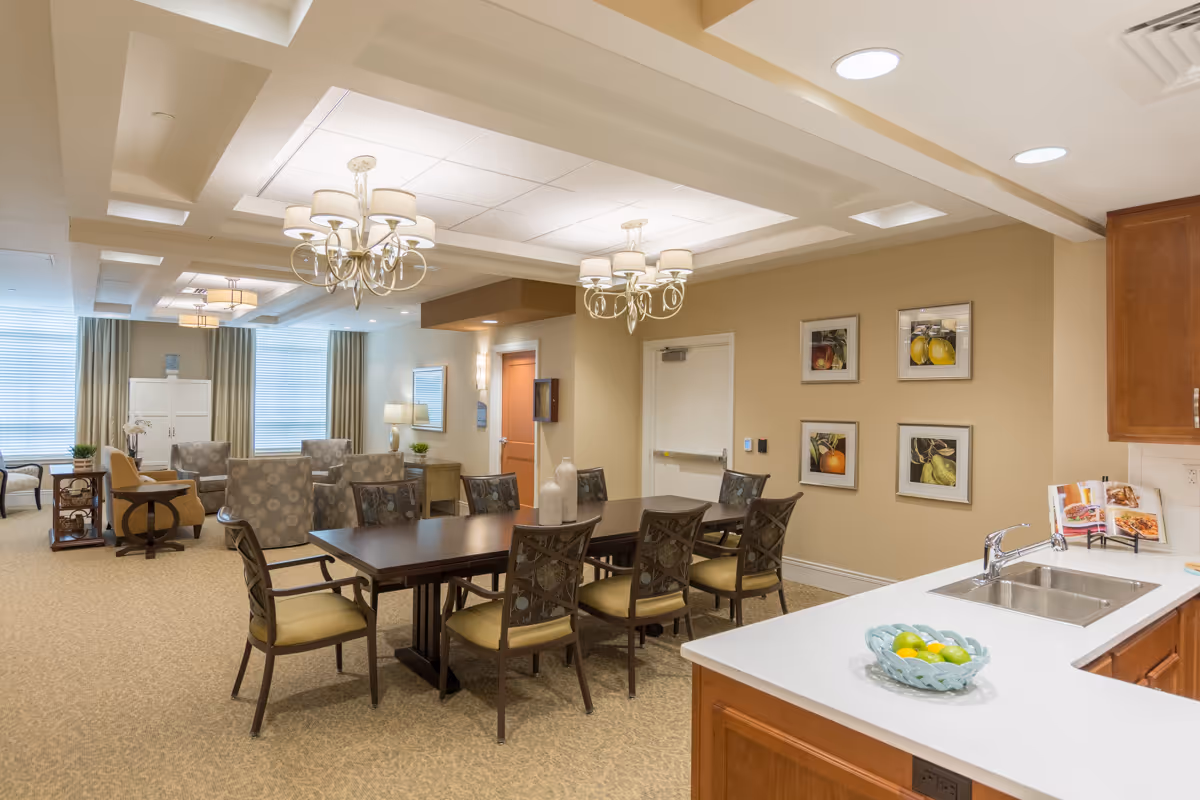 Open communal dining and seating area with a long wooden table and chairs, chandeliers, lounge seating by large windows, and a kitchenette countertop with a sink and fruit bowl.