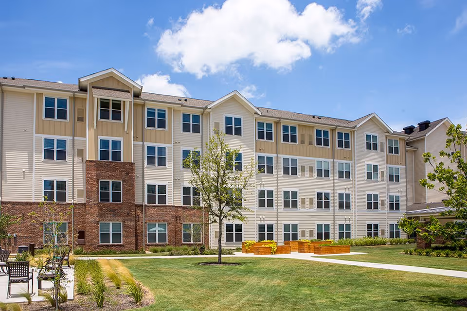 Exterior view of a multi-story senior living facility building with beige and brick facade, multiple windows, a well-maintained lawn, small trees, and garden beds under a partly cloudy blue sky.