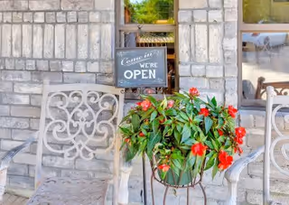 Two decorative metal chairs on a stone patio with a potted plant featuring red flowers placed between them. A small sign hanging on the window behind the chairs reads 'Come in! WE'RE OPEN'.