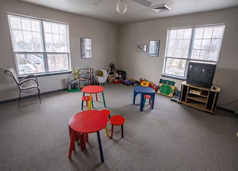 A bright playroom with two large windows letting in natural light. The room contains small colorful tables and stools, children's toys, a TV on a wooden stand, and a wicker chair near one window. The walls are light-colored and there are a few framed pictures hanging.