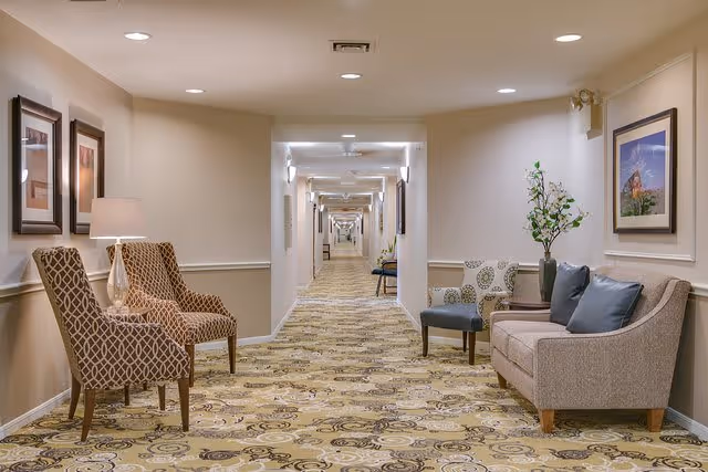 A long, well-lit hallway in a senior living facility with patterned carpet and beige walls. Along the hallway, there are several seating areas with upholstered chairs, a small sofa, a side table with a lamp, framed artwork on the walls, and a vase with flowers. The ceiling has recessed lighting.