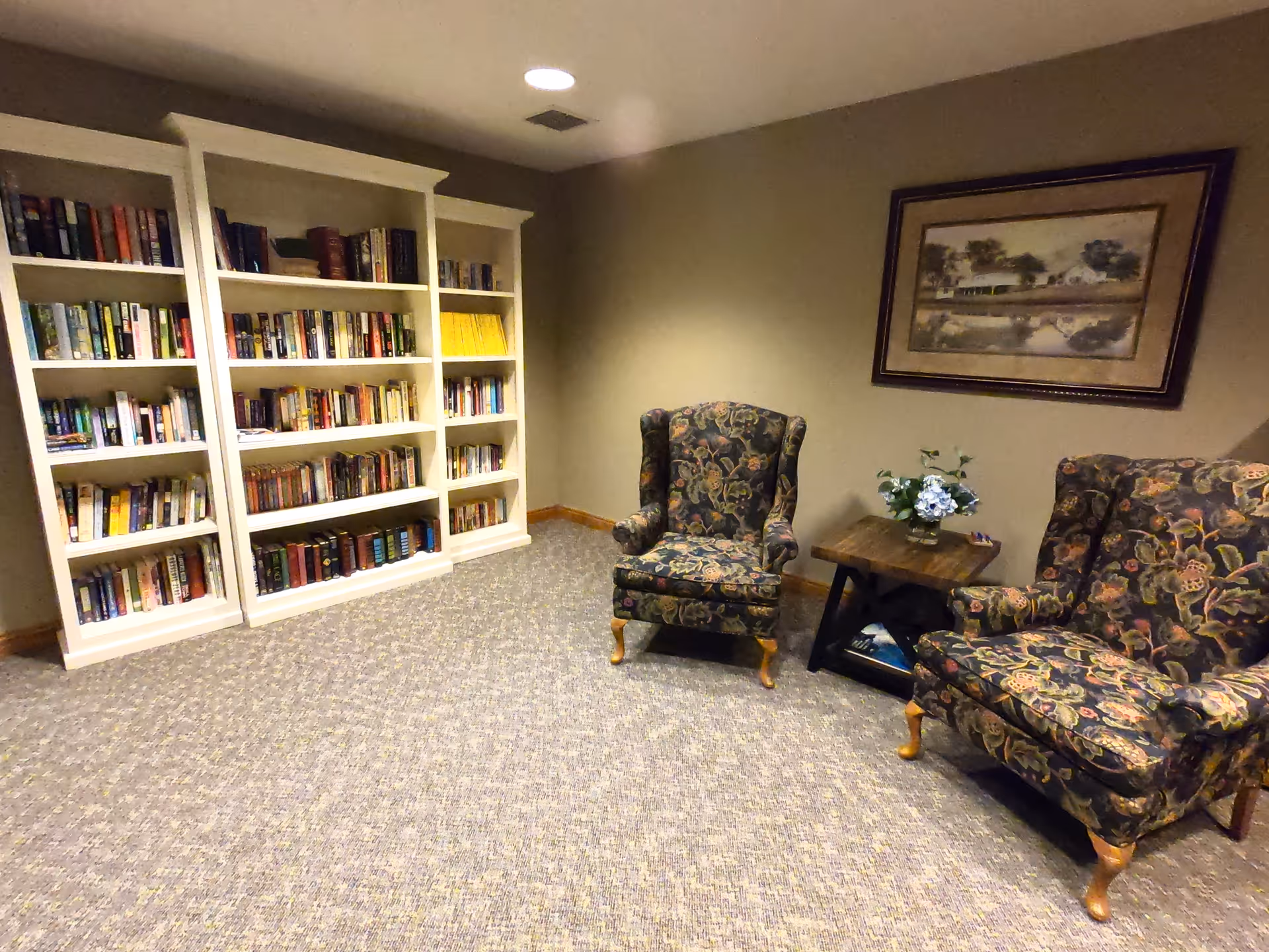 A cozy reading area in a senior living facility featuring two floral upholstered armchairs, a small wooden side table with a flower arrangement, a large framed painting on the wall, and a white bookshelf filled with books against a beige wall and carpeted floor.