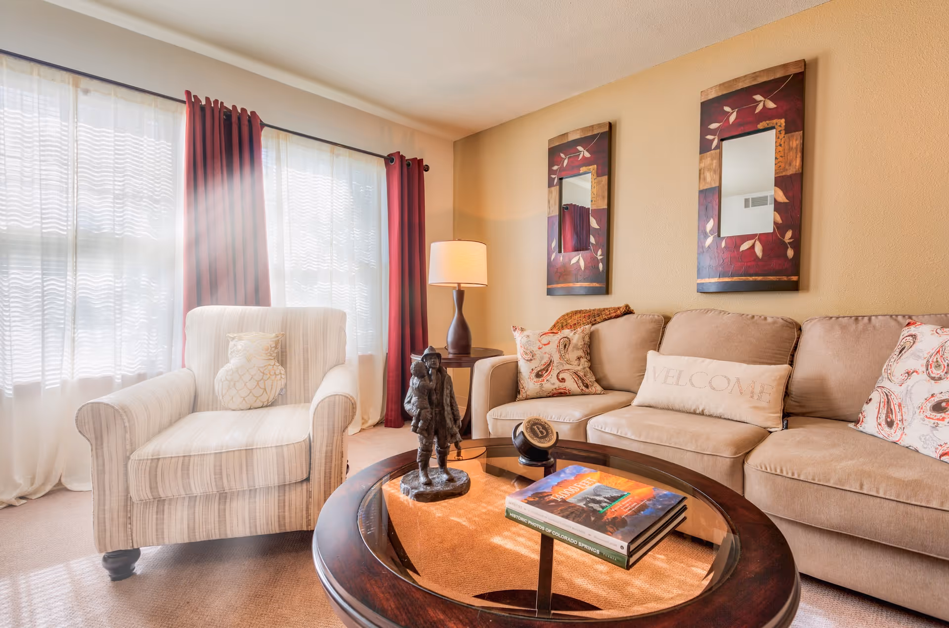 Sunlit living room with a striped armchair, beige sofa, round glass-top coffee table with books and decor, and two framed mirrors on the wall.