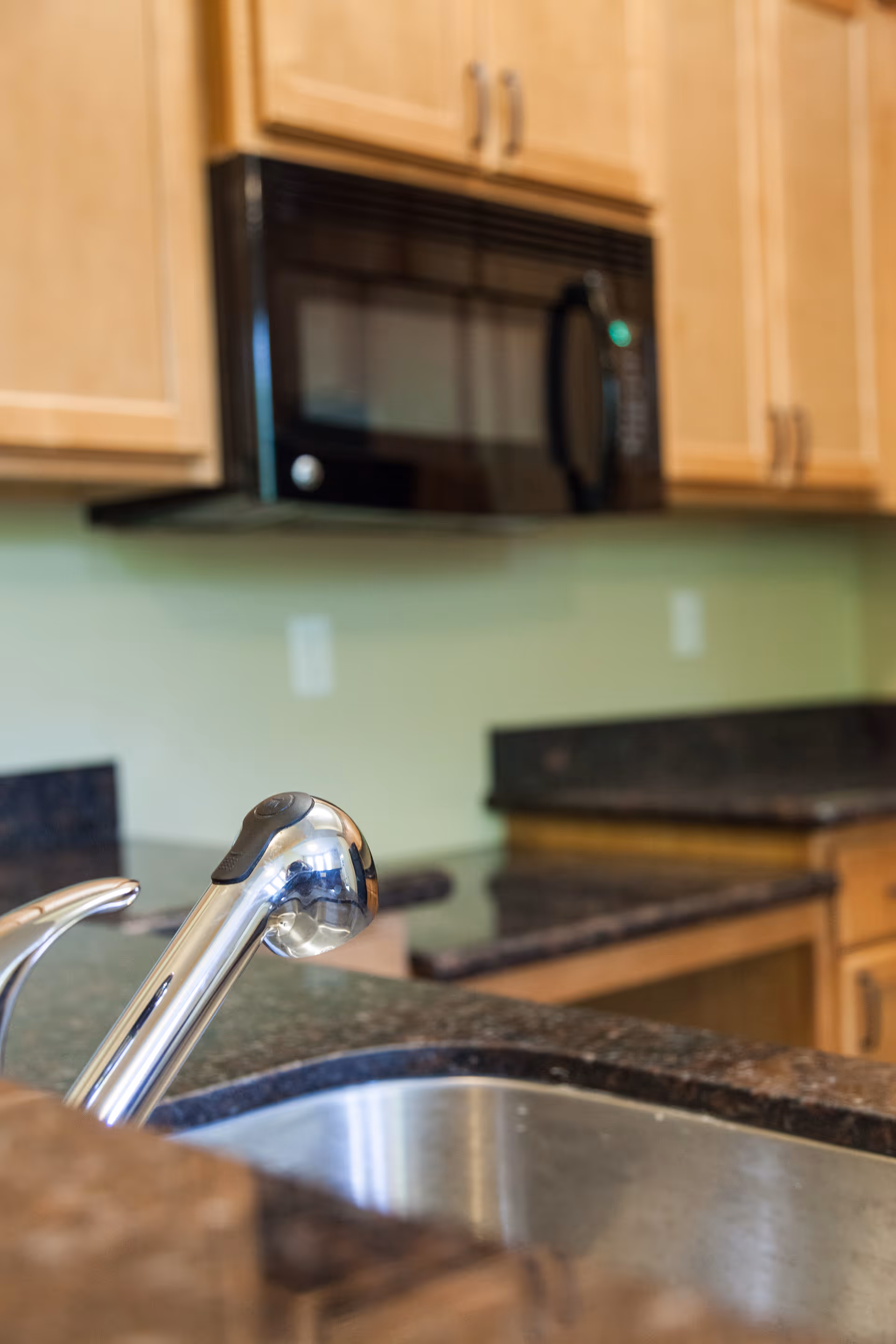 Close-up of a kitchen sink faucet with granite countertops, wooden cabinets, and a microwave in the background.