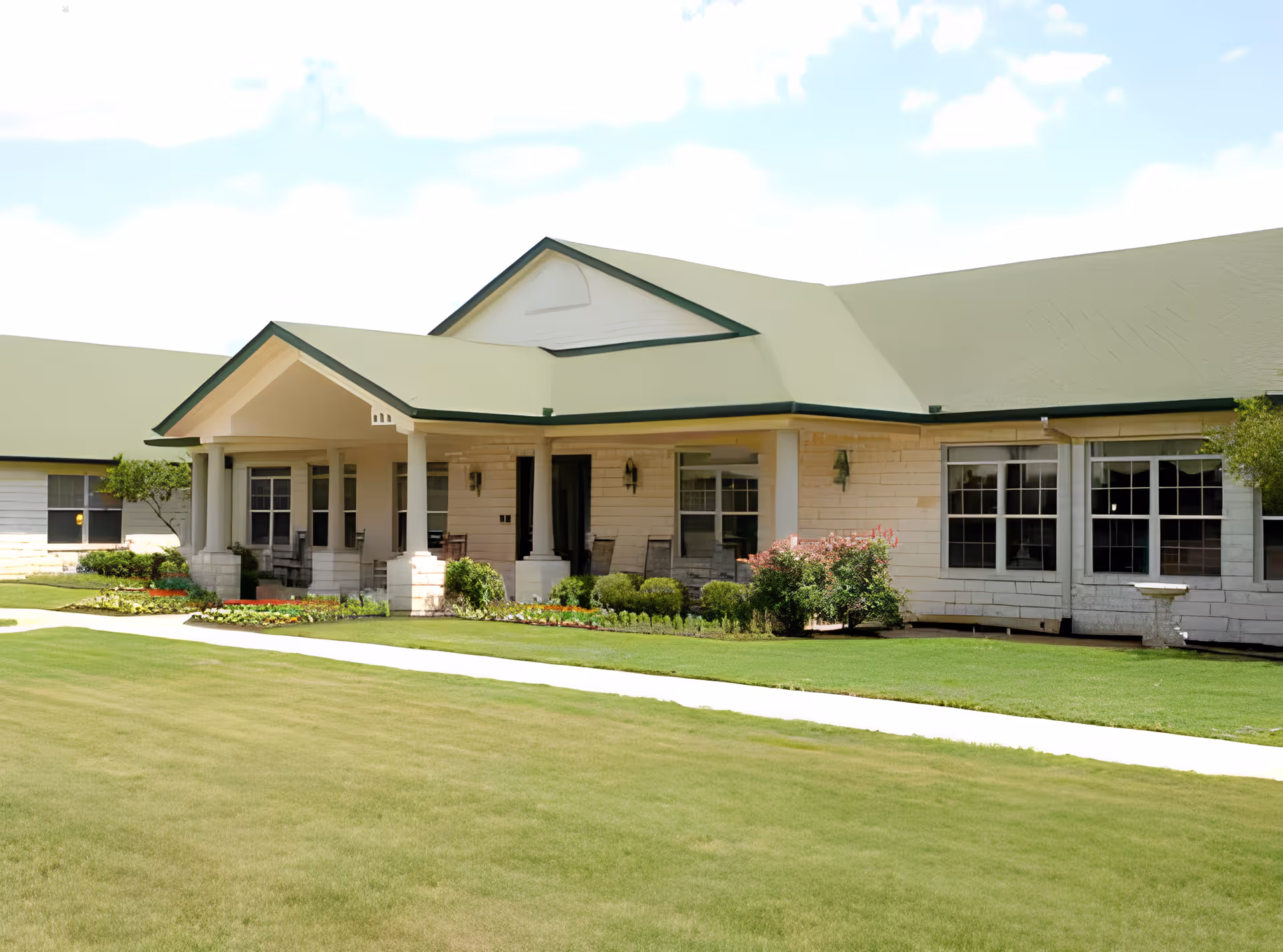 Front exterior of a single-story senior living building with a covered porch, green roof, and manicured lawn.