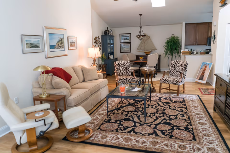 A cozy living room with a beige sofa, two zebra-patterned armchairs, a white recliner with matching footrest, a glass coffee table on a patterned rug, and a dining area with a round table and chairs in the background. The room features wooden flooring, framed artwork on the walls, a blue cabinet, a large green plant, and a model sailboat on a side table.
