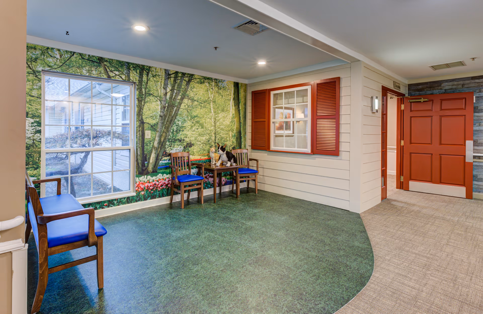 An interior common area designed to resemble an outdoor garden setting with a large wall mural of trees and flowers, a window with red shutters, two wooden chairs with blue cushions, a small table with decorative items, and a red door leading to another room.
