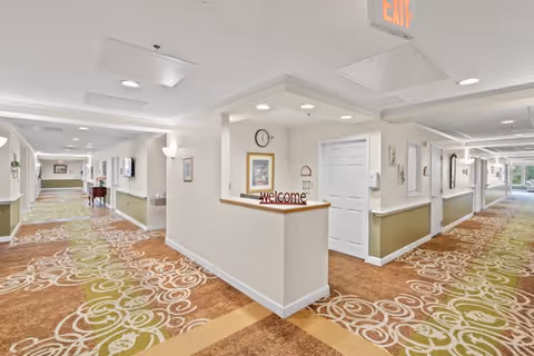 A bright, spacious hallway in a senior living facility with patterned carpet in shades of brown, green, and beige. The hallway features white walls with green wainscoting, recessed lighting, and several doors along the corridor. A small reception or welcome desk is visible in the center with a clock and framed pictures on the wall behind it. An exit sign is mounted on the ceiling.