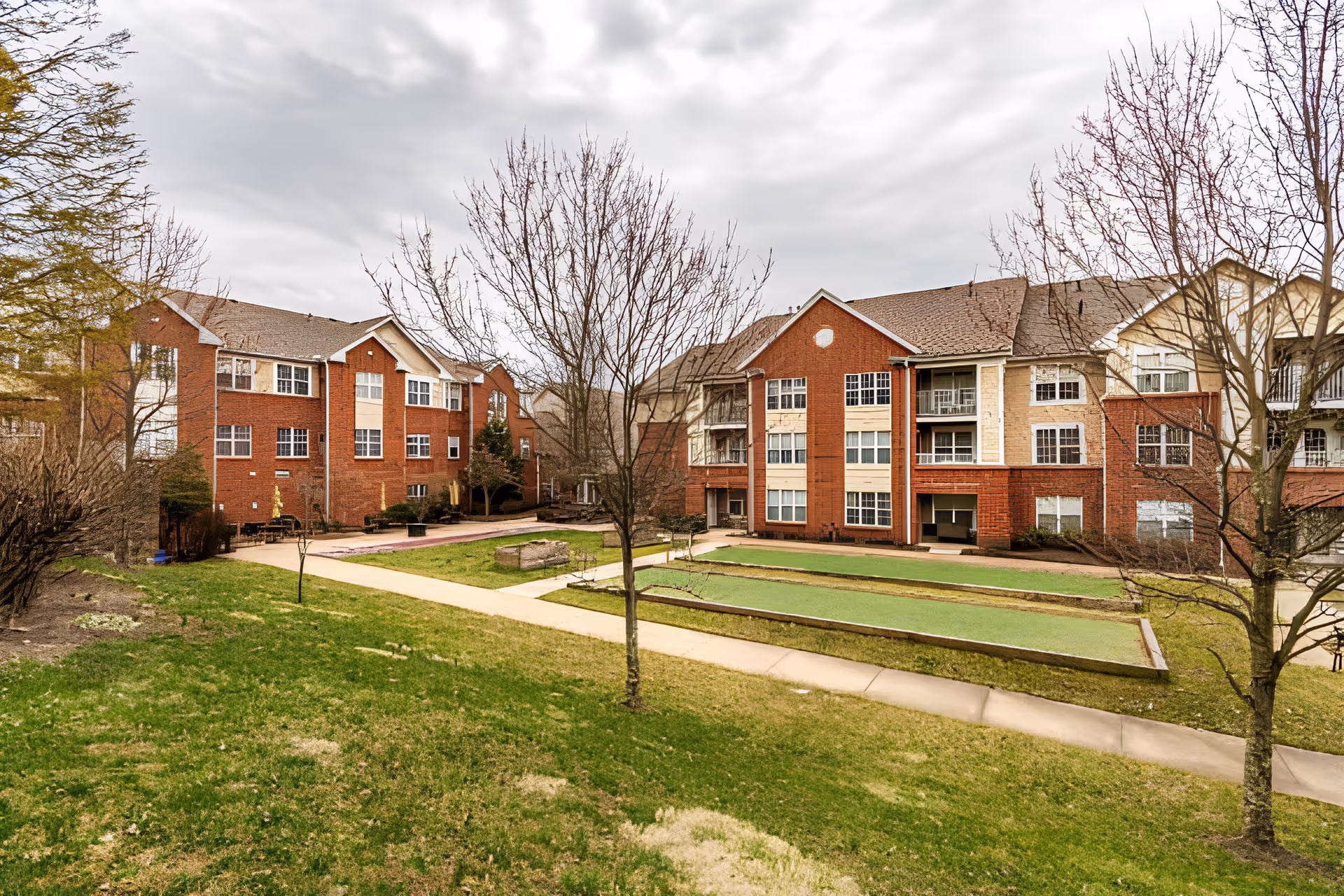 Courtyard with bocce-style lawns and walkways between red-brick, multi-story senior living buildings under an overcast sky.