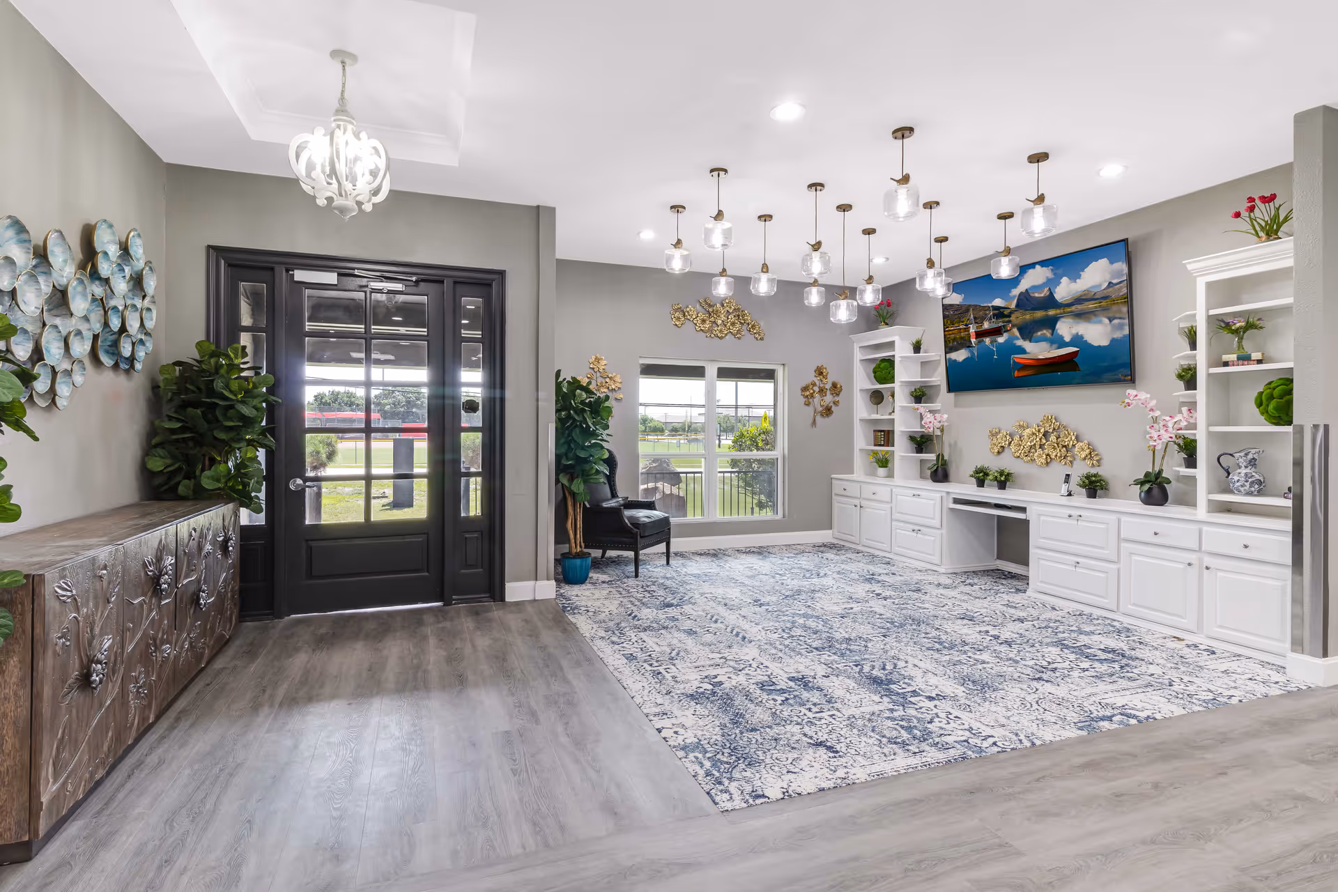 A bright and spacious interior room with a large patterned blue and white rug, white built-in cabinets and shelves decorated with plants and ornaments, a large flat-screen TV mounted on the wall displaying a scenic lake and mountain view, a black door with glass panels leading outside, and modern pendant lights hanging from the ceiling.