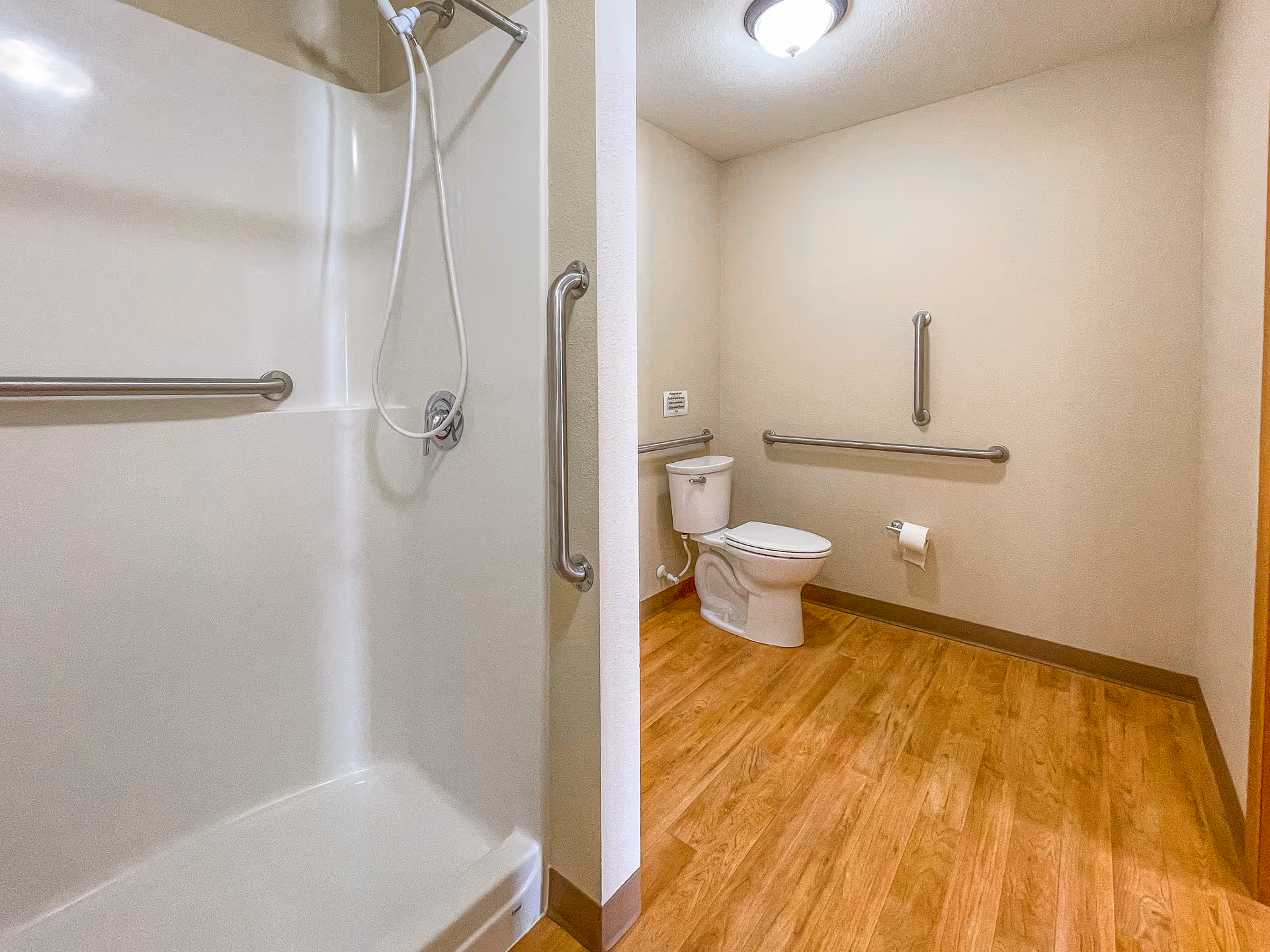 A spacious bathroom with a white shower area featuring a handheld showerhead and grab bars, and a toilet with grab bars on the walls. The floor is wood-patterned, and the walls are light beige with a ceiling light fixture.