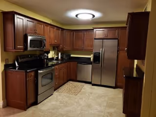 A kitchen with wooden cabinets, stainless steel appliances including a refrigerator, oven, microwave, and dishwasher. The floor is tiled, and there is a small rug in front of the oven. The walls are painted yellow and the ceiling light is circular and bright.