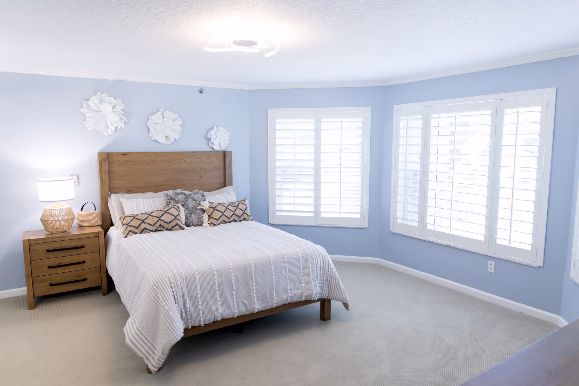 A bright and spacious bedroom with light blue walls and beige carpet. The room features a wooden bed frame with a white textured bedspread, decorative pillows, and a stuffed animal. There is a wooden nightstand with a lamp and a small basket. Three white decorative wall hangings are above the bed. Two large windows with white plantation shutters allow natural light to fill the room.