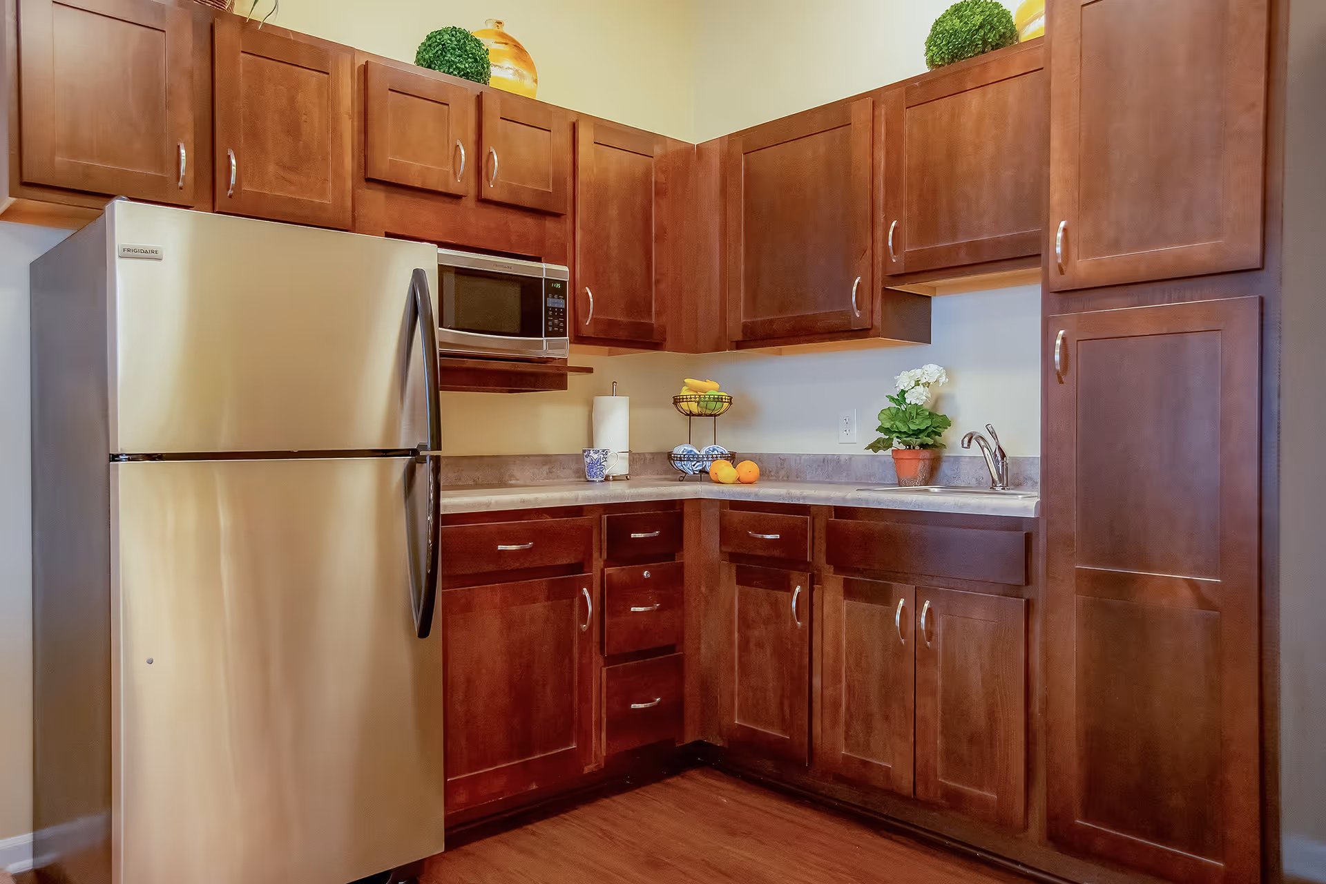 A kitchen with wooden cabinets, a stainless steel refrigerator, a microwave mounted above the counter, a paper towel holder, a fruit basket with bananas and oranges, a small potted plant with white flowers, and a sink with a faucet.