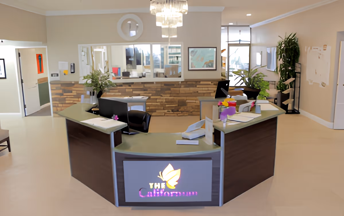 Reception area of The Californian facility featuring a modern curved front desk with a lit sign displaying the facility name and logo. The desk has papers, flowers, and office supplies on it. Behind the desk are chairs, plants, a wall with wood paneling, mirrors, and framed artwork. The space is well-lit with a chandelier and natural light from windows.