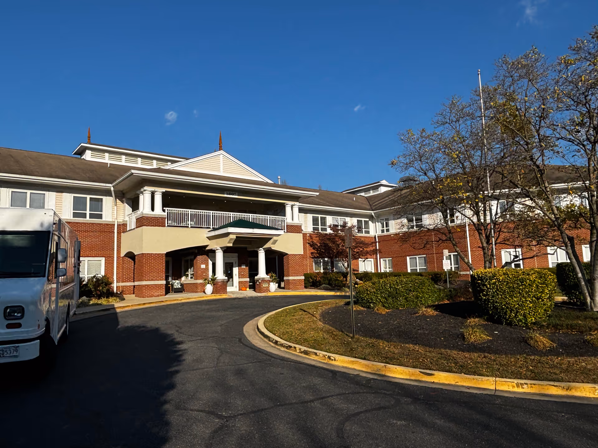 Exterior view of a senior living facility building with a curved driveway, landscaped bushes, and trees under a clear blue sky. The building has a brick and beige facade with white columns and a covered entrance.