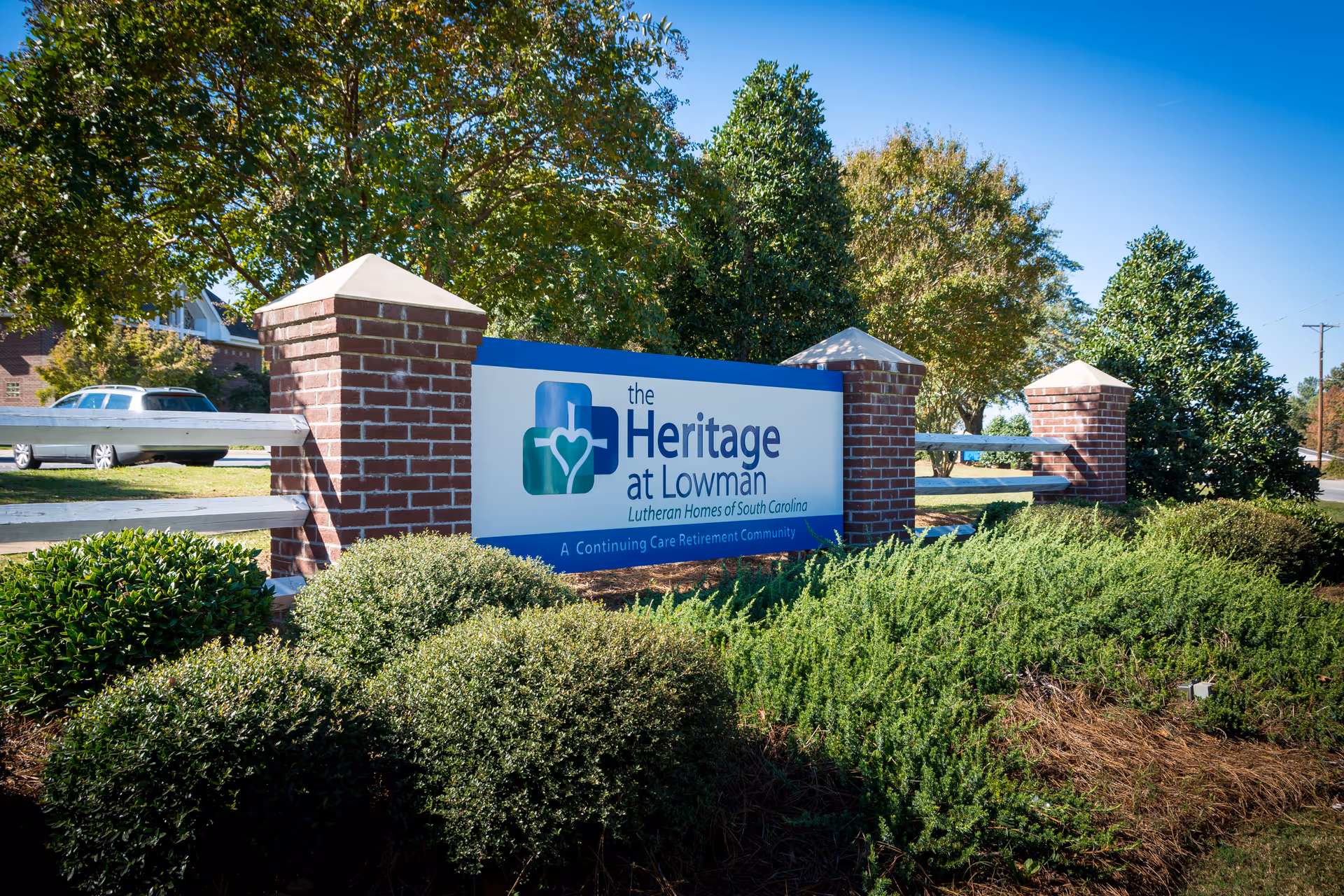 Outdoor view of a sign for The Heritage at Lowman, a continuing care retirement community, surrounded by green bushes and trees with a clear blue sky in the background.