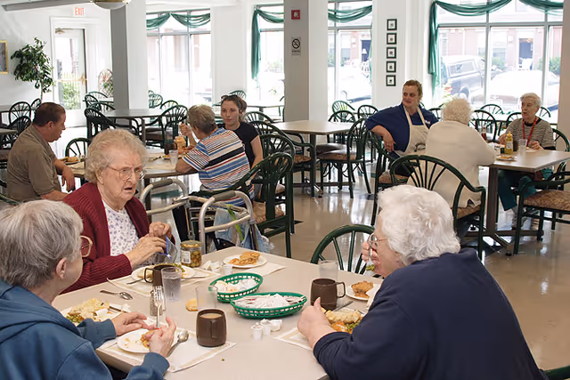Elderly residents eating and talking in a communal dining room with tables and green chairs.