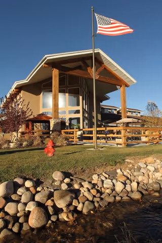 Exterior view of a building with large wooden beams supporting an overhanging roof, an American flag on a flagpole in front, a red fire hydrant on a grassy area, and a rocky edge bordering a small body of water under a clear blue sky.