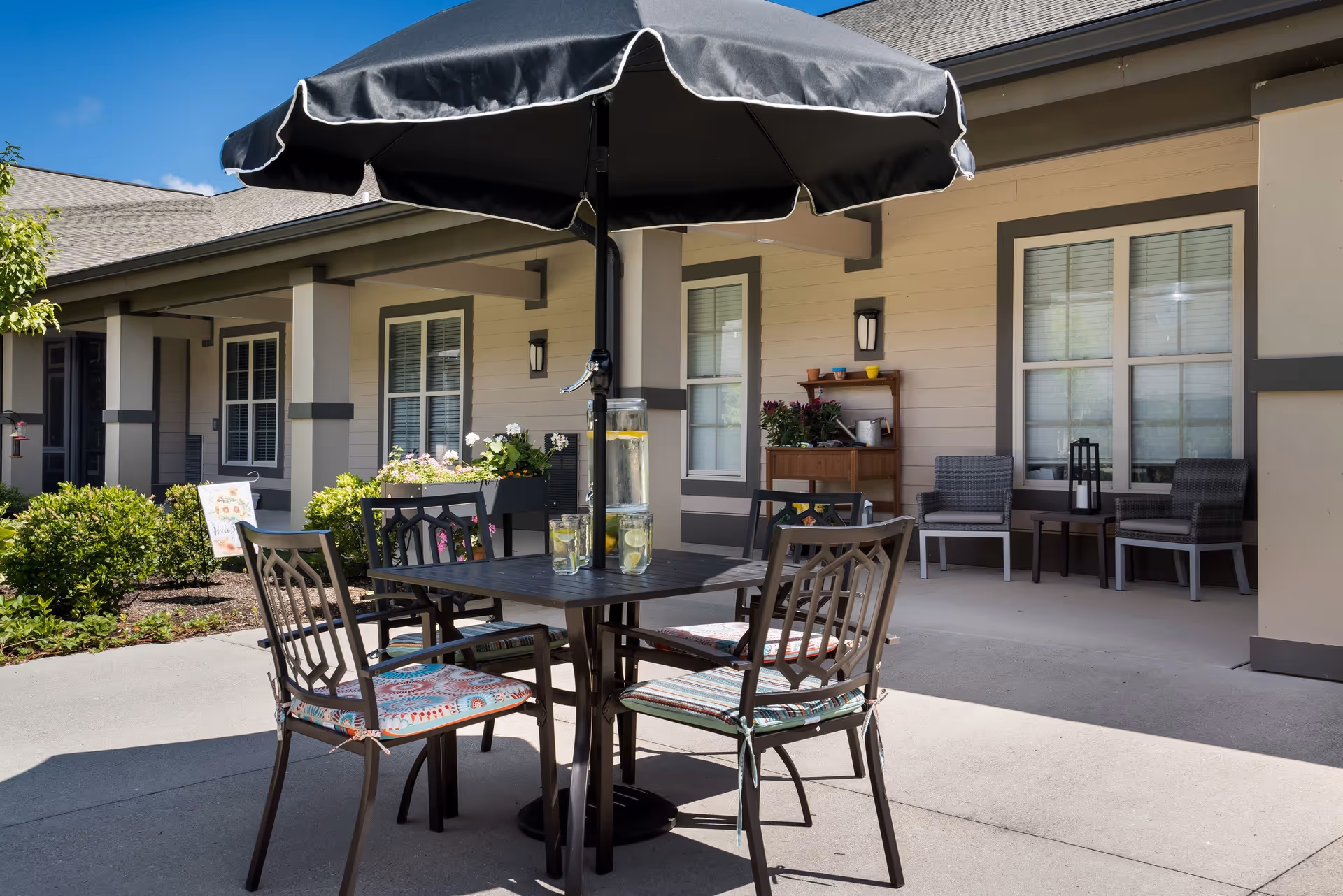 Outdoor patio area at Liberty Place Memory Care featuring a black umbrella over a square table with four chairs, each with patterned cushions. The table has a glass pitcher and glasses with lemon water. In the background, there are windows, a small wooden shelf with potted plants, two cushioned chairs with a small table between them, and some greenery along the building.
