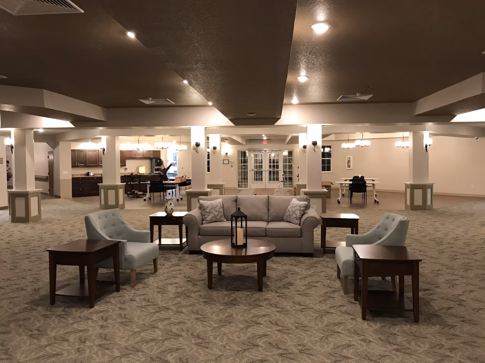 A spacious, well-lit common area in an assisted living facility with a beige sofa, two light blue armchairs, and several wooden side tables arranged on a patterned carpet. In the background, there is a kitchen area with dark wood cabinets and a person waving, as well as tables and chairs near large glass doors.