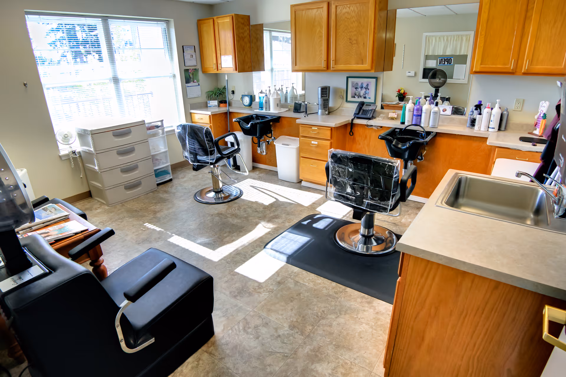 Bright salon-style room with two styling chairs, shampoo sinks, countertops and cabinets in a senior living facility.