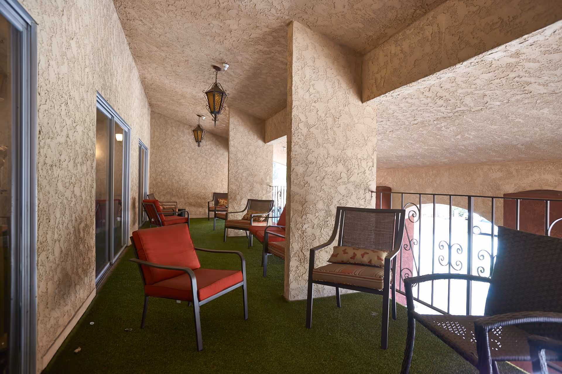 Covered outdoor patio area with textured beige walls and ceiling, green artificial grass flooring, and several chairs with red and patterned cushions arranged along the space. Decorative hanging lanterns are suspended from the ceiling, and a wrought iron railing borders the open side overlooking a parking area.