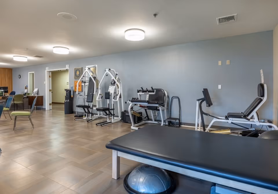 A spacious exercise room in a senior living facility with various workout equipment including weight machines, a padded exercise bench, and chairs. The room has light blue walls, wooden flooring, and ceiling lights.