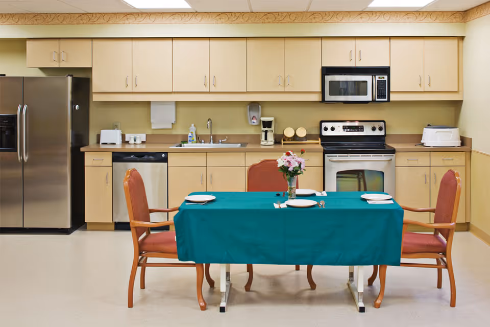 A clean kitchen area with beige cabinets, a stainless steel refrigerator, dishwasher, stove, microwave, and toaster. In front of the kitchen counter is a table covered with a green tablecloth, set with four plates, utensils, and a vase with flowers. Four wooden chairs with red cushions surround the table.