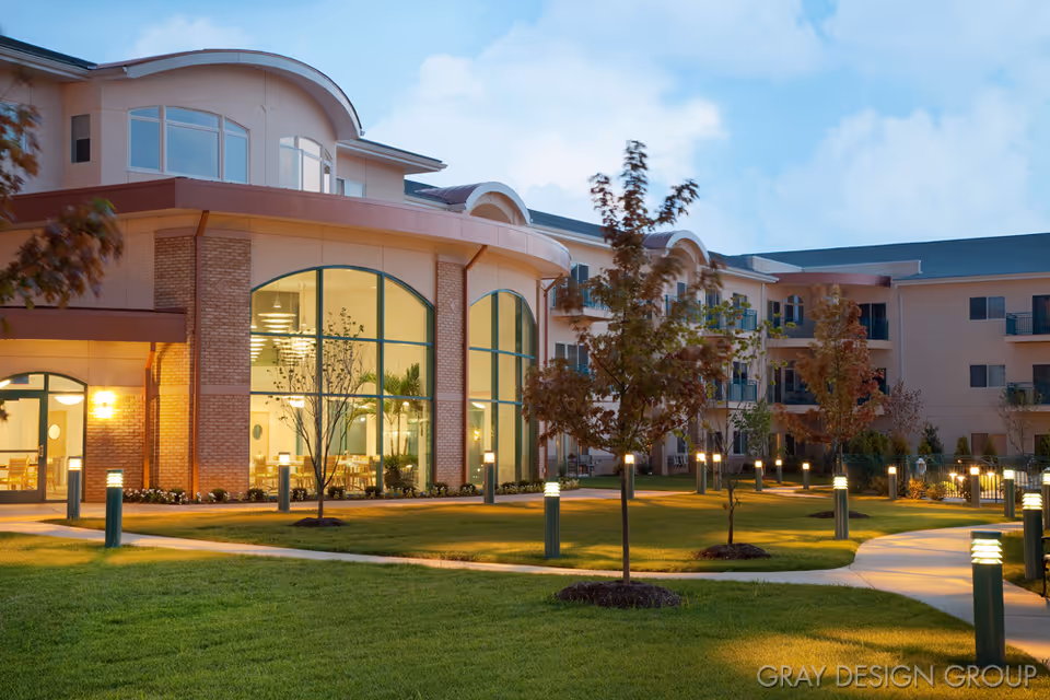 Exterior view of a senior living facility at dusk with well-lit pathways, manicured lawns, young trees, and large windows showing a dining area inside.