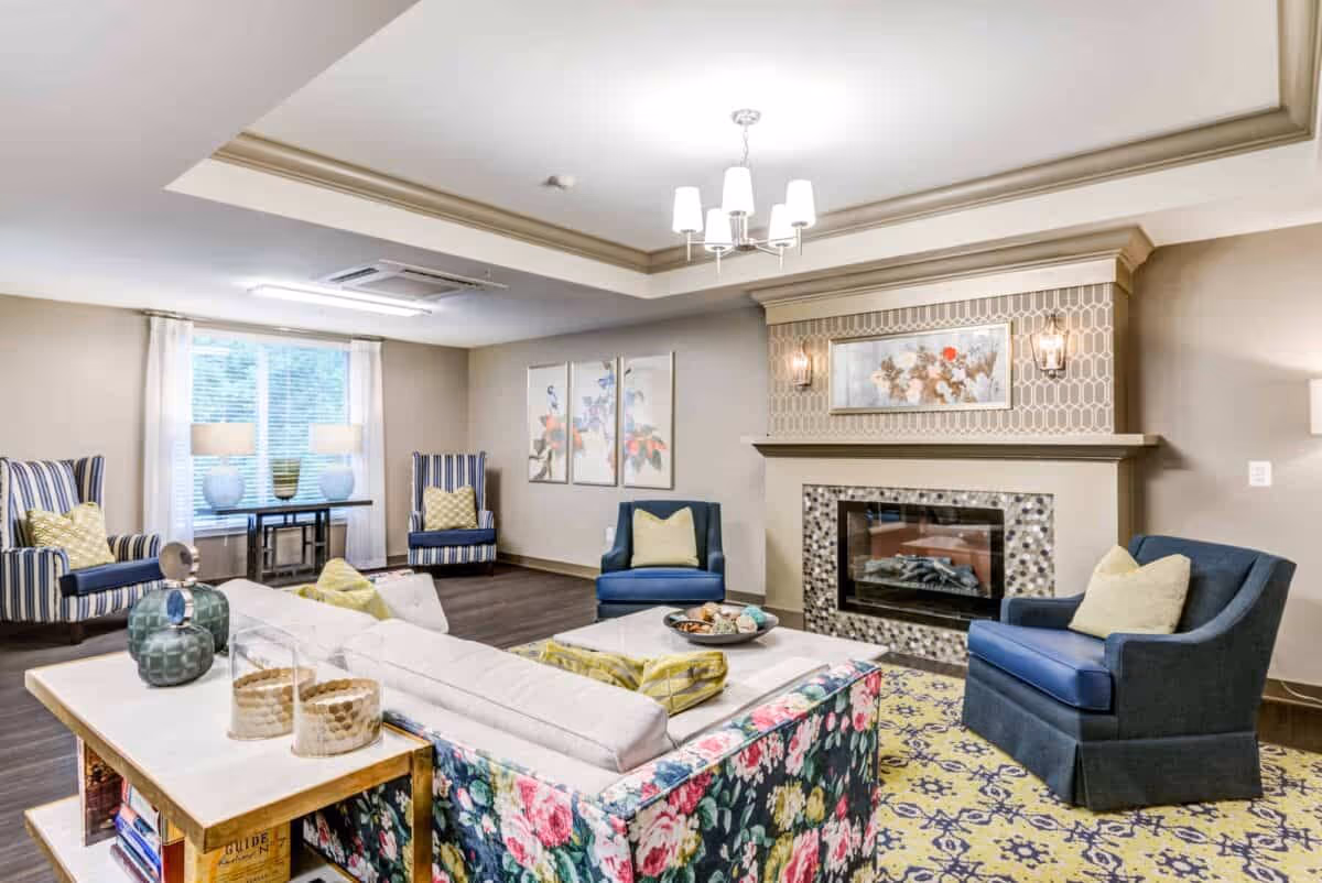 A bright and spacious living room with a floral-patterned sofa, two blue armchairs with yellow pillows, and two striped armchairs near a window with white curtains. The room features a decorative fireplace with a mosaic tile surround and a framed floral artwork above it. A yellow and blue patterned rug covers the floor, and a chandelier hangs from the ceiling.