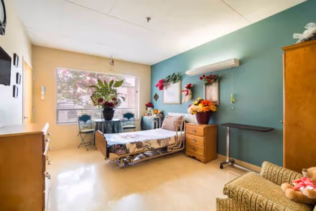 A bright and cozy senior living bedroom with a hospital-style bed covered in floral bedding, a wooden dresser, a small table with two chairs near a large window, and various flower arrangements and decorations on the walls and furniture. There is also a comfortable armchair with a teddy bear on it.