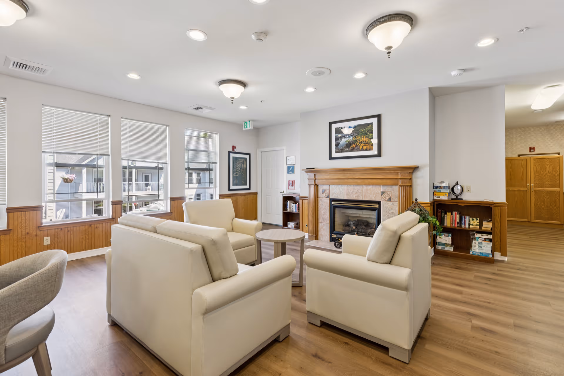 A bright and cozy living room area in a senior living facility with four cream-colored armchairs arranged around a small round wooden table. The room features large windows with blinds, a wooden floor, and a fireplace with a wooden mantle and a framed landscape picture above it. There are bookshelves with books and games, and the walls are painted white with wooden paneling halfway up.