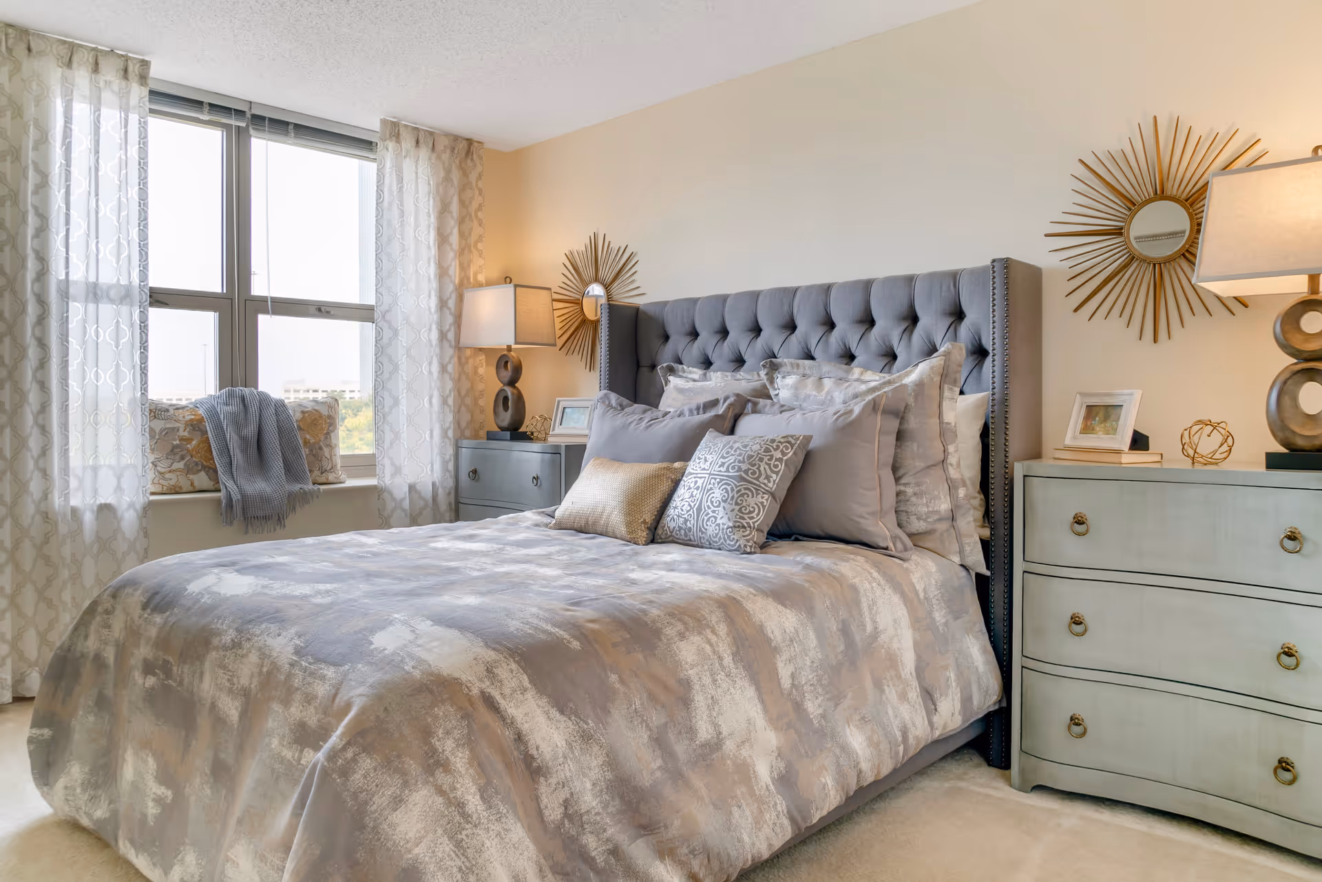 A cozy bedroom with a large bed featuring a tufted gray headboard and multiple decorative pillows. The bed is covered with a patterned comforter in shades of gray and beige. On either side of the bed are matching light green nightstands with lamps and decorative items. Two sunburst mirrors hang on the wall above the nightstands. A window with sheer patterned curtains lets in natural light, and a cushioned window seat with pillows and a throw blanket is visible.