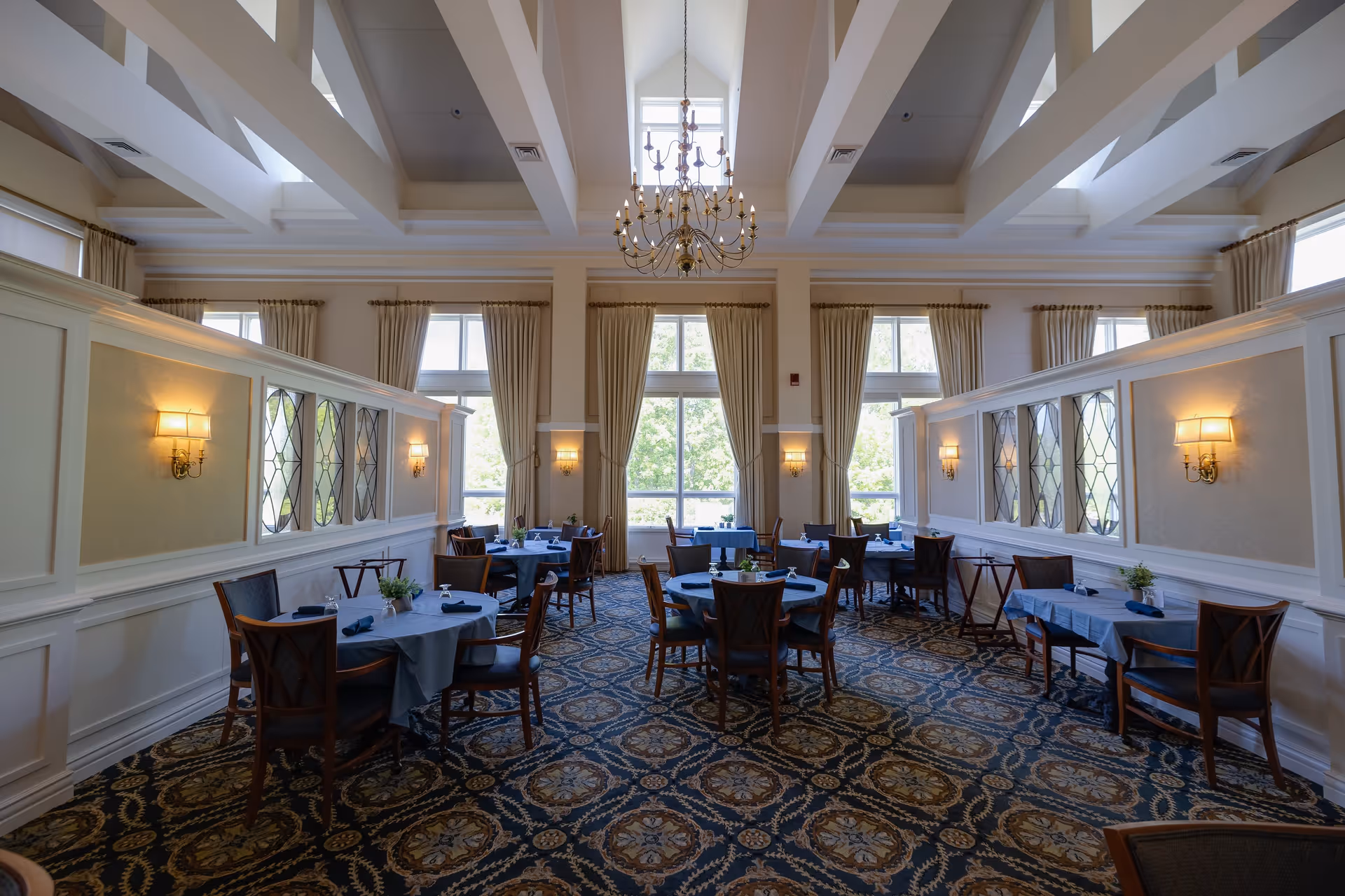 A bright and elegant dining room with multiple round tables covered with blue tablecloths and set with napkins and glassware. The room features large windows with beige curtains, a patterned carpet, wall sconces, and a chandelier hanging from a high ceiling with exposed beams.