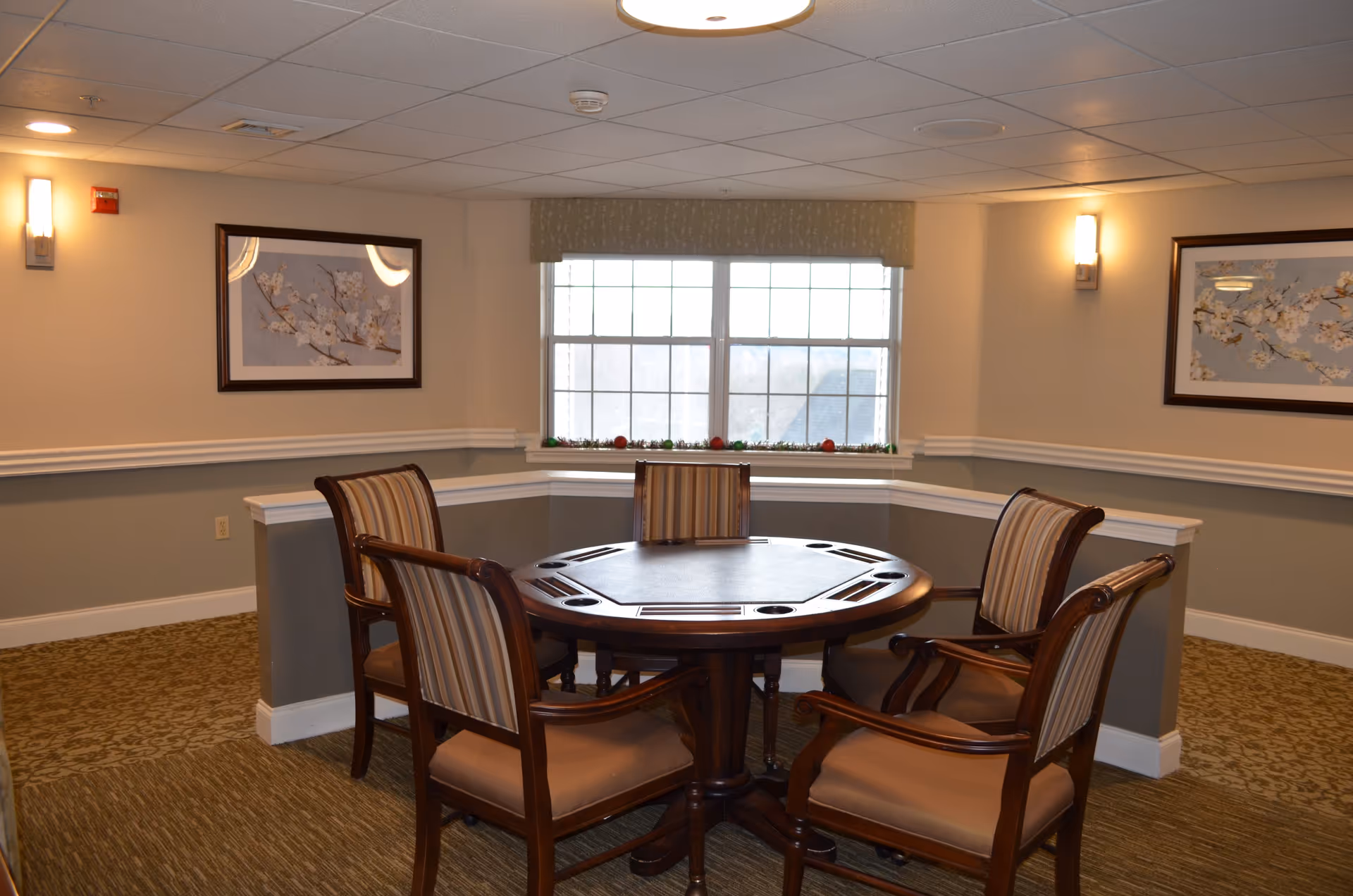 A cozy interior room with a round wooden table surrounded by four cushioned chairs with striped upholstery. The room has beige walls with white trim, two framed floral paintings, a window with a valance, and soft lighting from wall sconces.